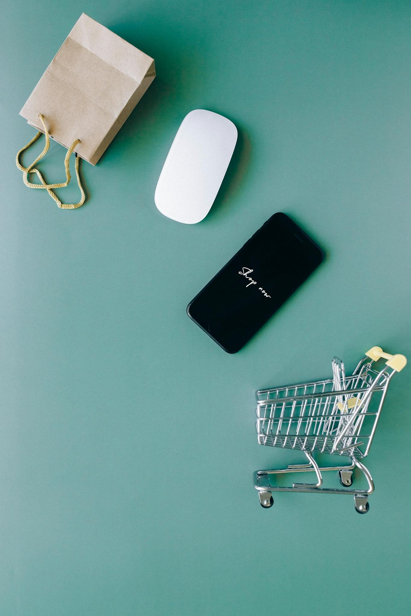 Top view of a smartphone, mouse, paper bag, and mini shopping cart on a green background, symbolizing e-commerce.